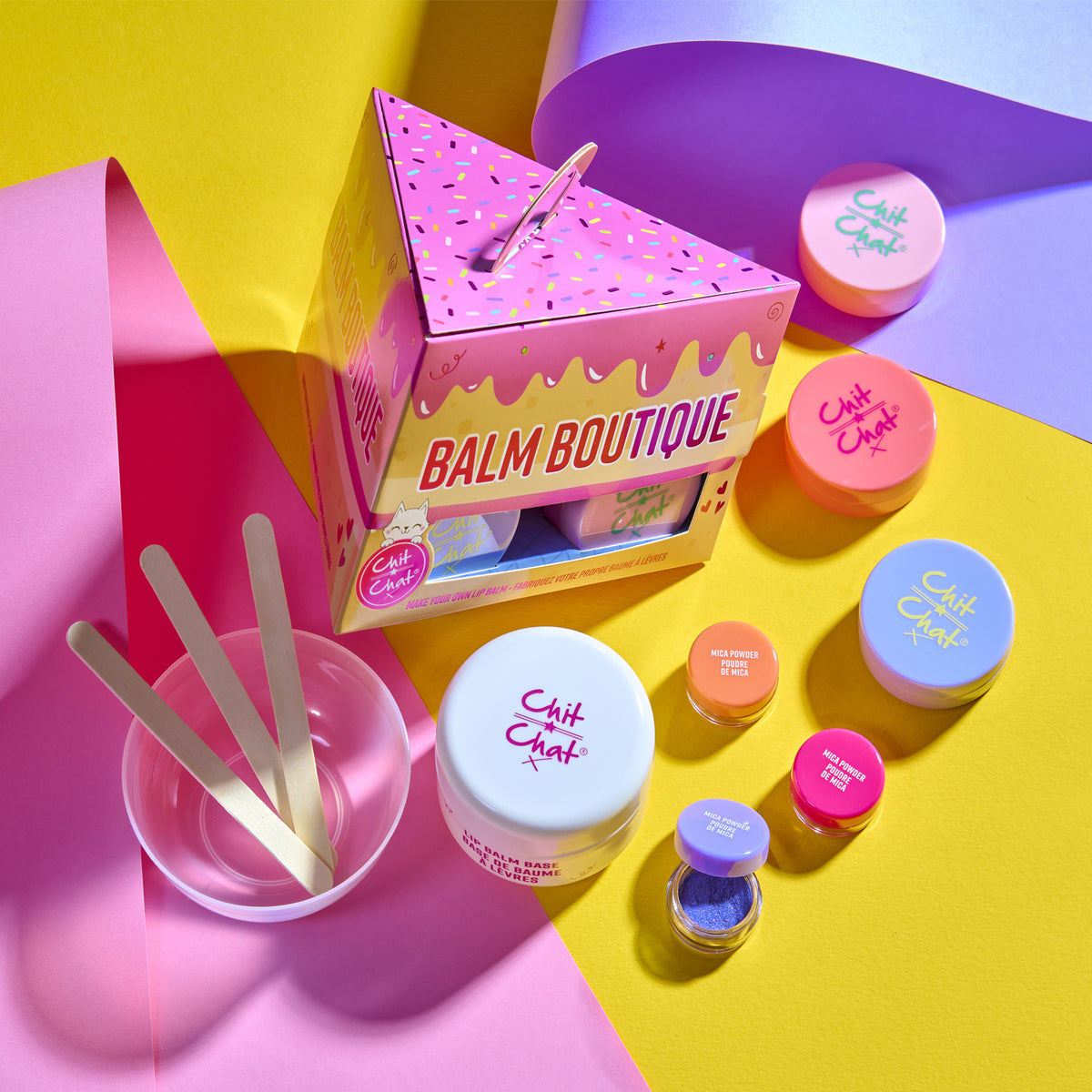 A colourful lip balm making kit set arranged on a bright multi-coloured background. The set includes a triangular pink box labeled 'Balm Boutique,' several round tubs with pastel lids, wooden spatulas in a clear cup, and small cosmetic containers.