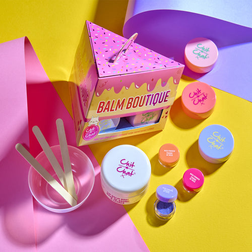 A colourful lip balm making kit set arranged on a bright multi-coloured background. The set includes a triangular pink box labeled 'Balm Boutique,' several round tubs with pastel lids, wooden spatulas in a clear cup, and small cosmetic containers.