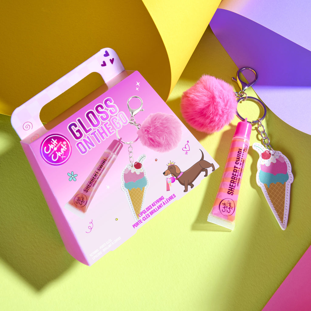 A pink gift set box labeled 'Gloss on the Go' with a lip gloss tube and pink pom-pom keychain and ice cream keychain, arranged on a bright multicolour background.