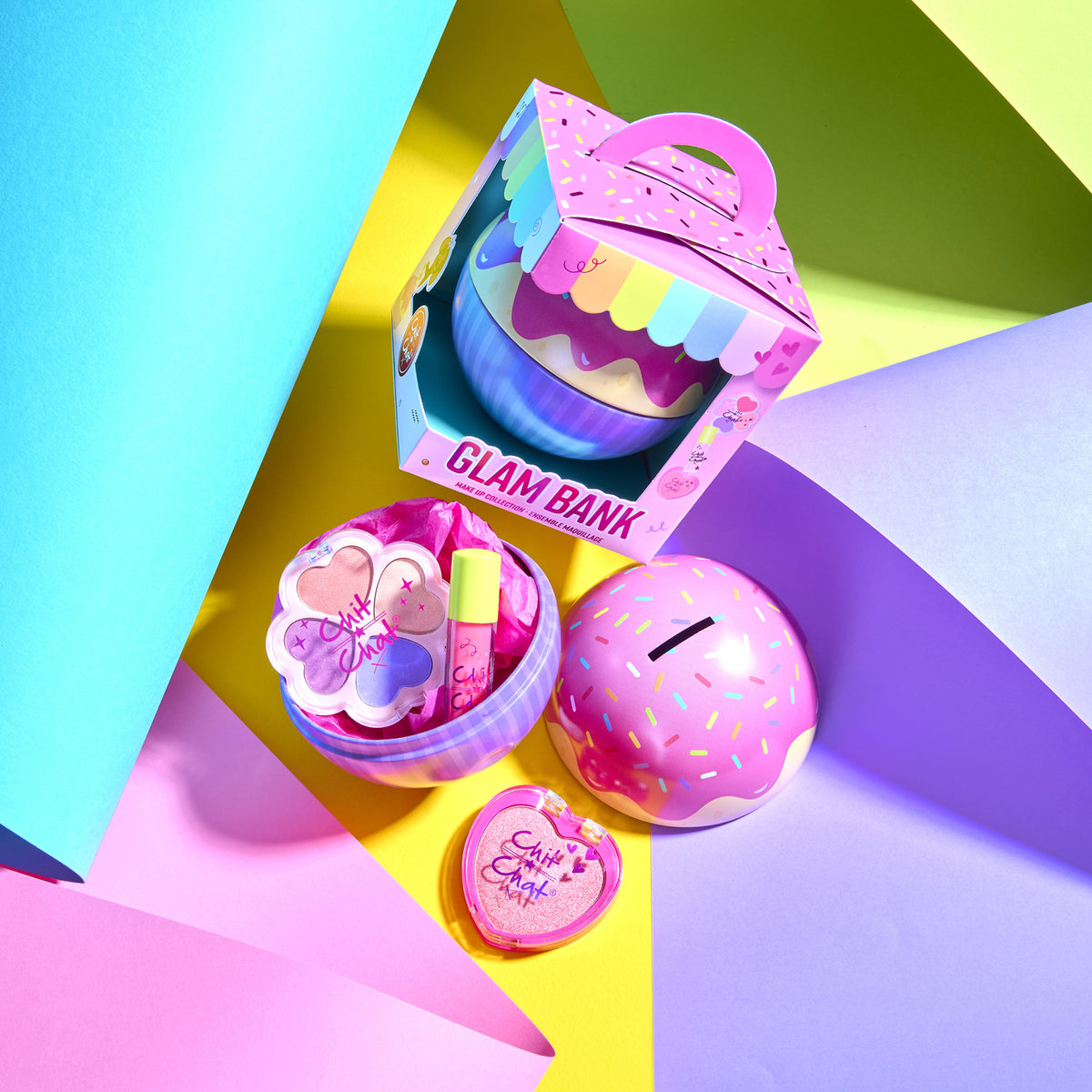 A colourful cosmetic gift set arranged on a geometric pastel background. The set includes a pink cupcake-themed money tin in a packaged box labelled 'GLAM BANK,' a pink lip product tube, a flower shaped 4 colour eyeshadow compact, and a heart-shaped blusher.