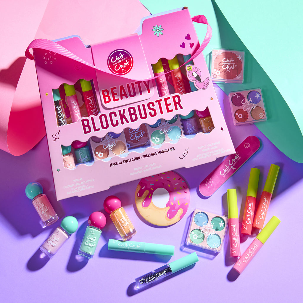 A pink Beauty Blockbuster makeup collection gift box displayed with various colourful makeup items including lip glosses, nail polishes, mascaras and face palettes on a vibrant purple and teal background.