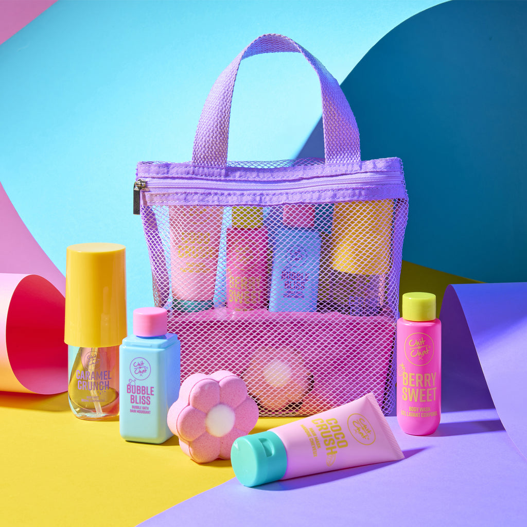 A colourful toiletry set arranged on a bright, geometric studio background. The set includes a lavender mesh zip bag with four colourful bottles, and a flower-shaped bath bomb.