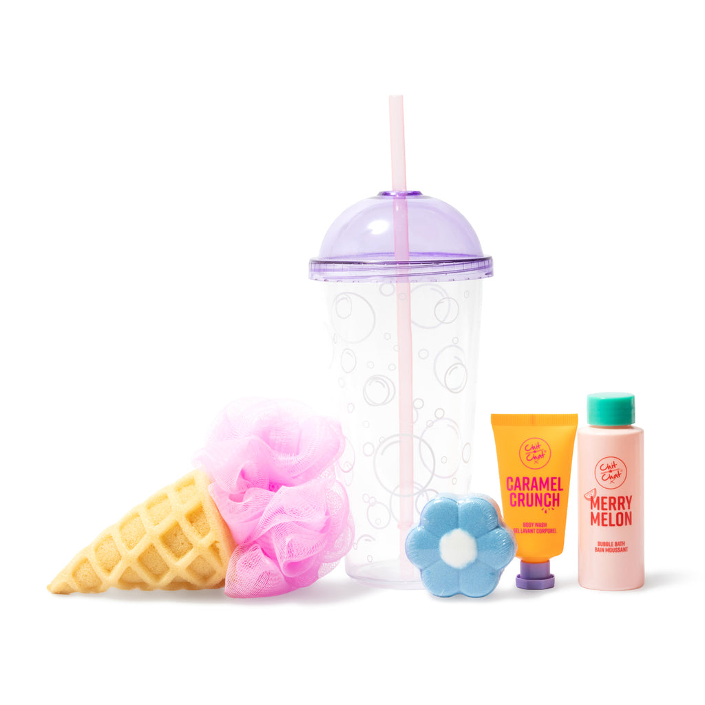 A bubble plastic cup with a purple lid and straw filled with a pink mesh loofah, a yellow waffle cone sponge, a blue flower-shaped bath bomb, an orange tube of Caramel Crunch body wash, and a pink bottle of Merry Melon bubble bath arranged on a white background.