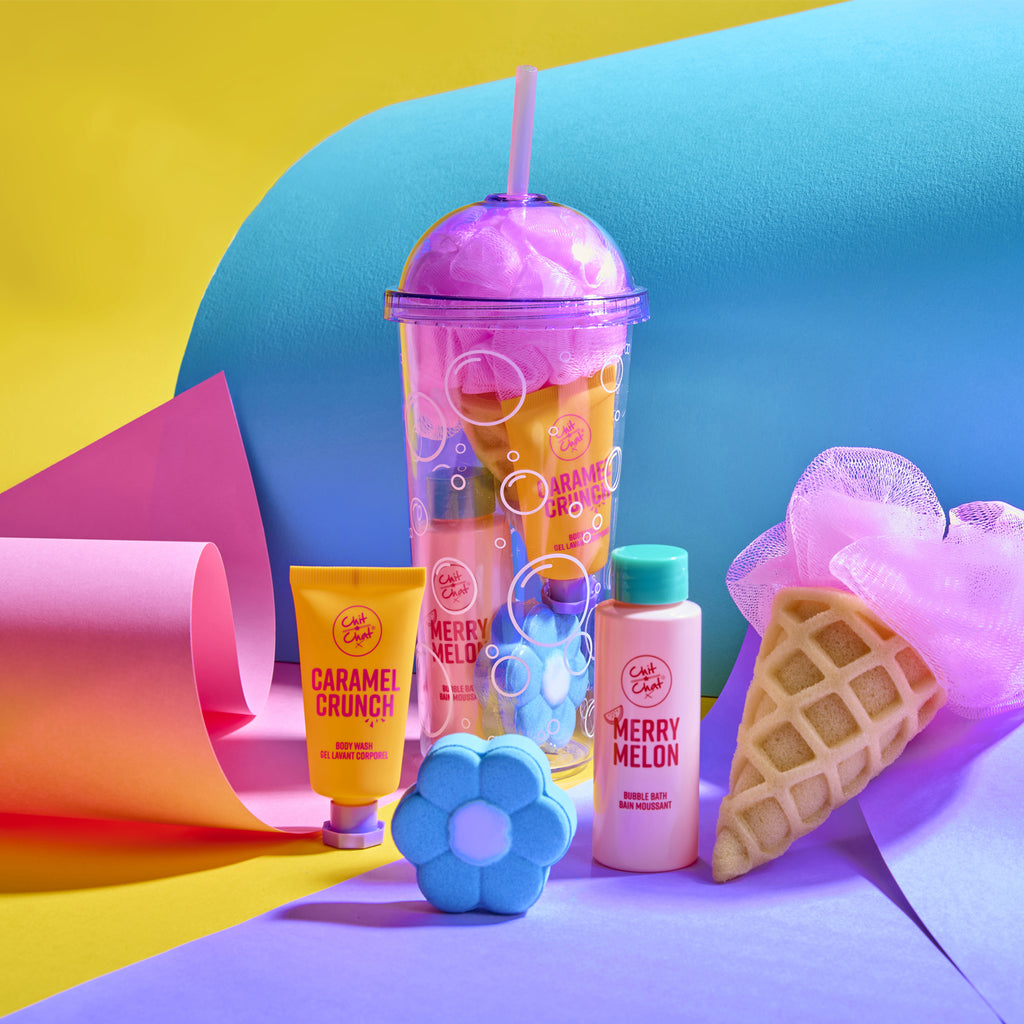 A colourful bath and body gift set styled  on a bright studio background. The set includes a translucent purple tumbler with a bubble design, a yellow tube labelled Caramel Crunch, a pink bottle labelled Merry Melon, a blue flower-shaped fizzer, and a waffle cone sponge and pink shower loofah.