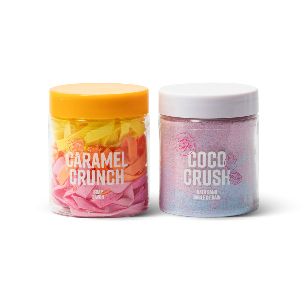 Two tubs of bath products. A clear jar with a yellow lid labelled 'Caramel Crunch' containing yellow, orange, pink soap string. A clear jar with a white lid labelled 'Coco Crush' containing light purple-blue bath sand. 