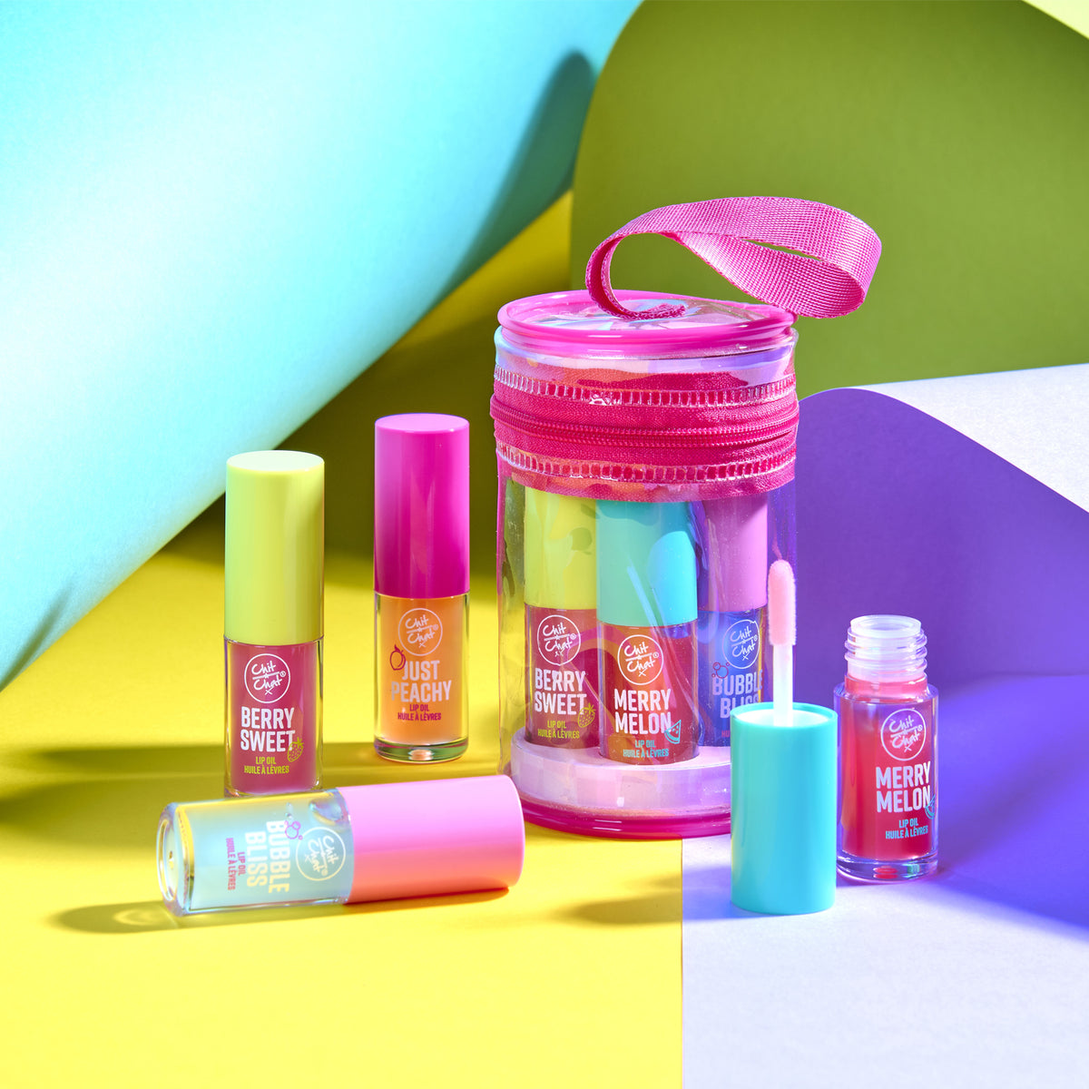 A set of lip oils arranged on a colourful geometric studio background. The collection includes four lip oils in pink shades, baby blue, orange, along with a clear cylindrical case with a pink zipper and pink ribbon handle.  