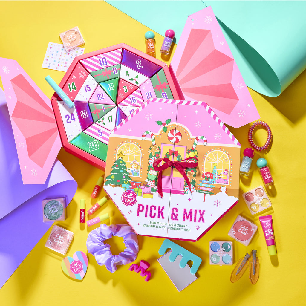 A colourful cosmetic advent calendar arranged on a bright yellow surface with pastel paper rolls in the background. The main product is a pink 'Pick & Mix' advent calendar with a Christmas decorated house illustration, surrounded by assorted beauty items and accessories. 